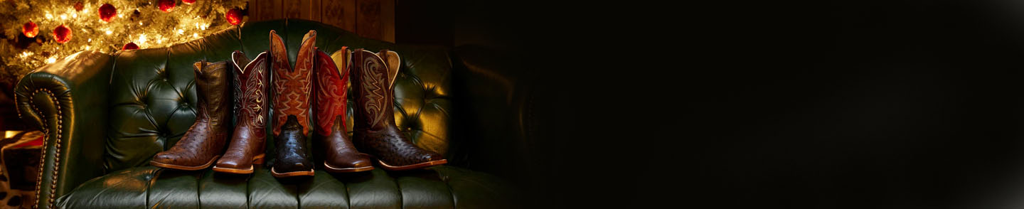 Imagen de una fila de botas Tony Lama sobre un sofá verde con un árbol de Navidad al fondo.